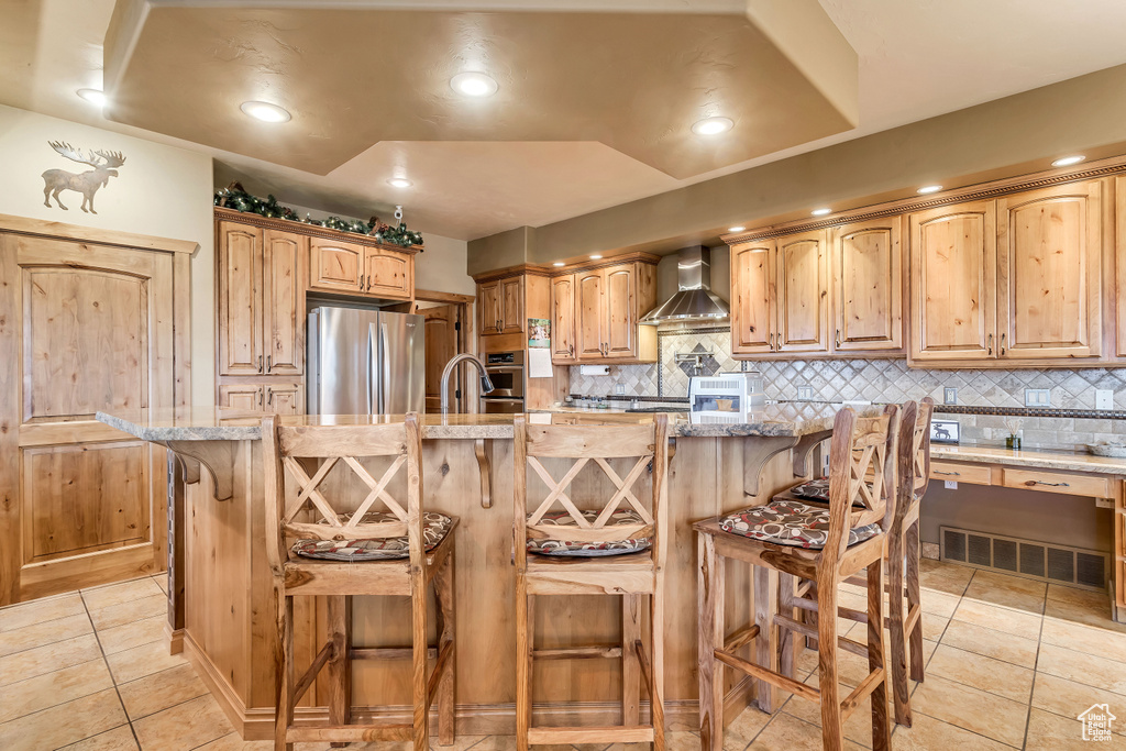 Kitchen with light tile patterned flooring, a breakfast bar area, wall chimney exhaust hood, appliances with stainless steel finishes, and a center island with sink