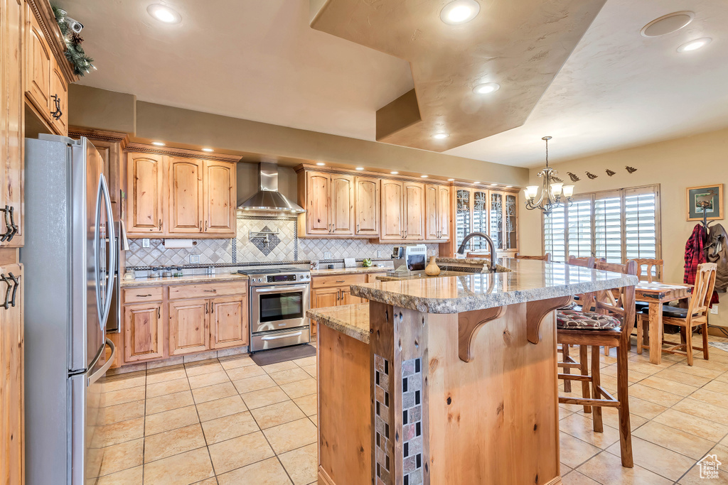 Kitchen featuring a kitchen bar, stainless steel appliances, a center island with sink, hanging light fixtures, and wall chimney exhaust hood