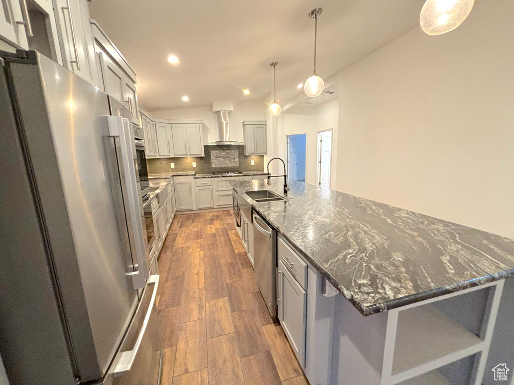 Kitchen with stainless steel appliances, wood-type flooring, wall chimney exhaust hood, and a center island with sink