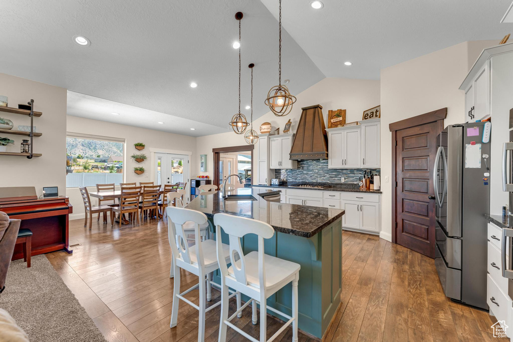 Kitchen featuring decorative light fixtures, wood-type flooring, a center island with sink, stainless steel refrigerator, and lofted ceiling