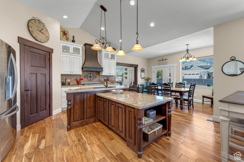 Kitchen with stainless steel appliances, hanging light fixtures, white cabinets, custom range hood, and light hardwood / wood-style flooring