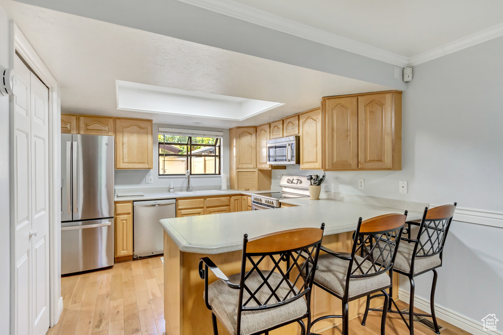 Kitchen with appliances with stainless steel finishes, kitchen peninsula, light hardwood / wood-style floors, light brown cabinetry, and ornamental molding