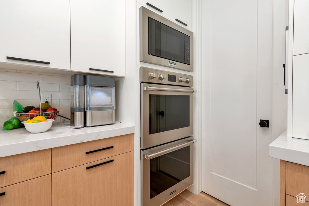 Kitchen with black microwave, backsplash, light wood-type flooring, white cabinetry, and double oven