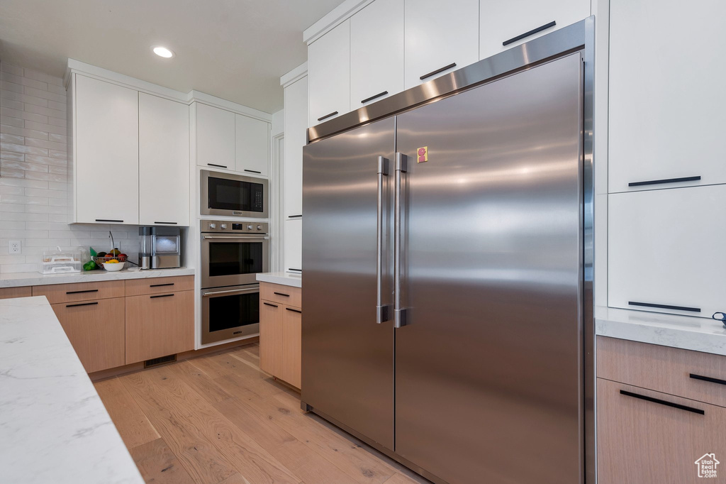 Kitchen with white cabinets, light hardwood / wood-style floors, light stone countertops, built in appliances, and decorative backsplash