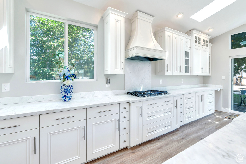 Kitchen with stainless steel gas cooktop, white cabinets, light stone countertops, custom exhaust hood, and lofted ceiling with skylight