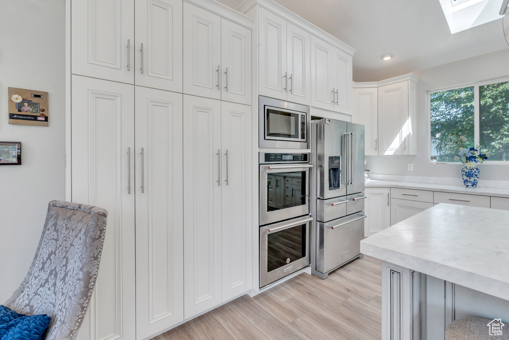 Kitchen with a skylight, white cabinetry, light wood-type flooring, stainless steel appliances, and light stone countertops