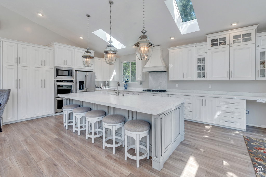 Kitchen featuring a skylight, white cabinetry, stainless steel appliances, light hardwood / wood-style floors, and custom exhaust hood