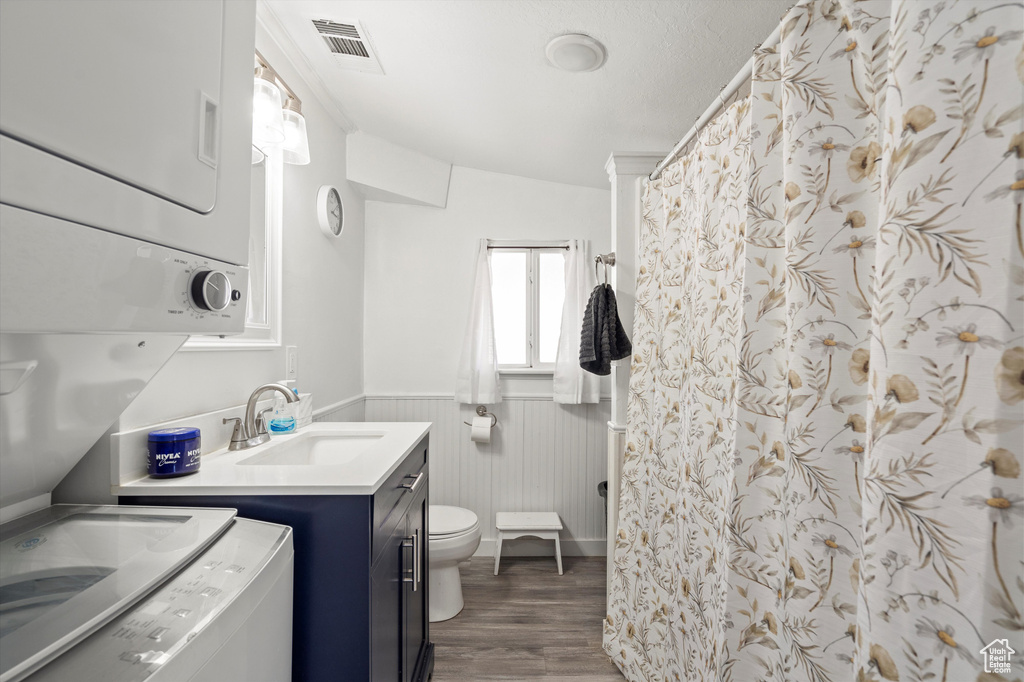 Bathroom with stacked washer / drying machine, toilet, vanity, wood-type flooring, and vaulted ceiling