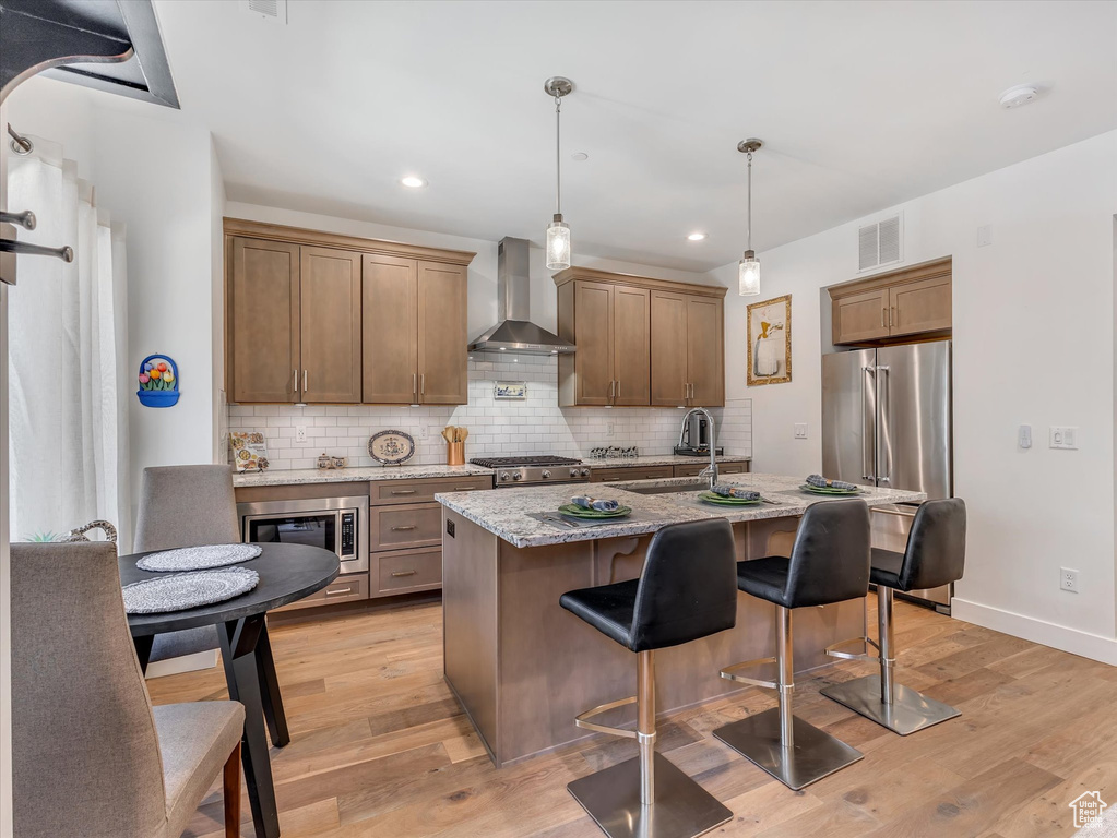 Kitchen with light hardwood / wood-style flooring, wall chimney range hood, backsplash, appliances with stainless steel finishes, and pendant lighting