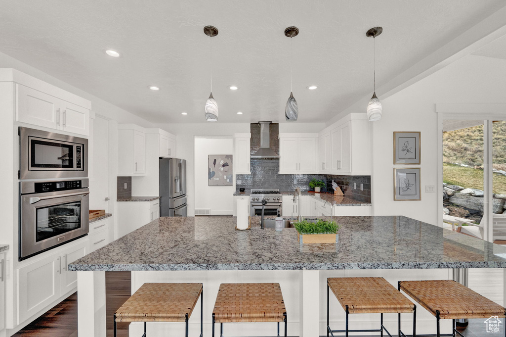 Kitchen with wall chimney range hood, dark wood-type flooring, a large island, appliances with stainless steel finishes, and decorative backsplash