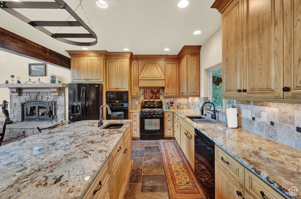 Kitchen with light stone countertops, black appliances, custom exhaust hood, a stone fireplace, and sink