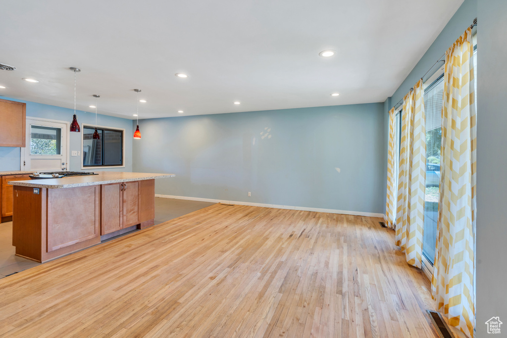 Kitchen with light wood-type flooring, a kitchen island, hanging light fixtures, stainless steel gas stovetop, and light stone counters