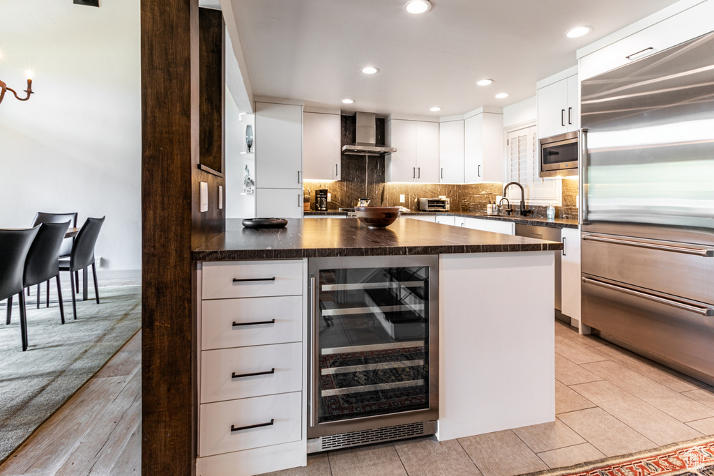 Kitchen featuring white cabinetry, wall chimney range hood, backsplash, beverage cooler, and built in appliances