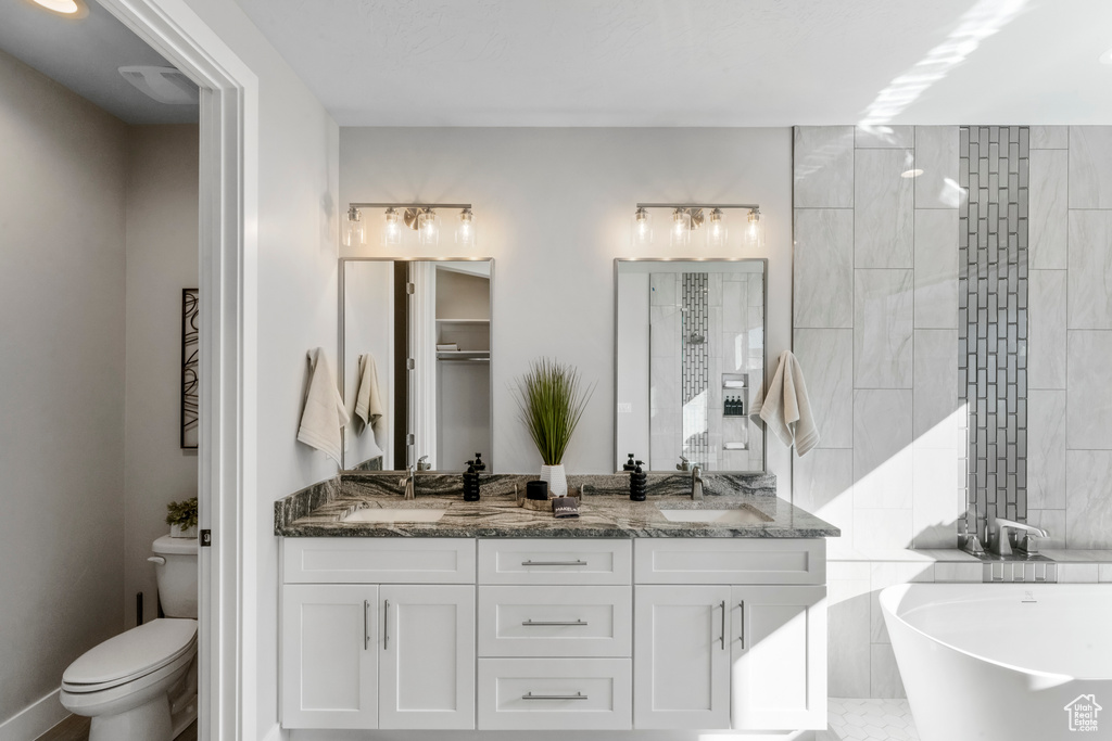 Bathroom featuring tile walls, a tub to relax in, toilet, and dual bowl vanity