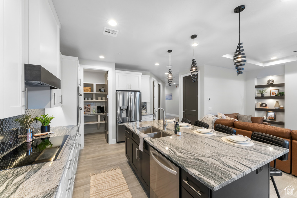 Kitchen featuring stainless steel appliances, white cabinets, an island with sink, wall chimney exhaust hood, and light hardwood / wood-style flooring