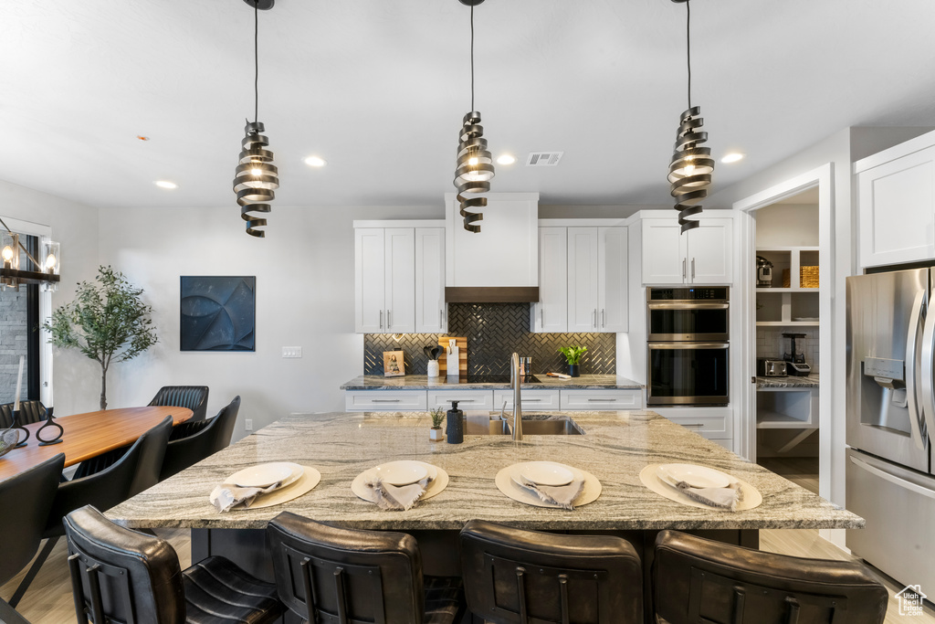 Kitchen featuring backsplash, light stone counters, an island with sink, stainless steel appliances, and a kitchen breakfast bar