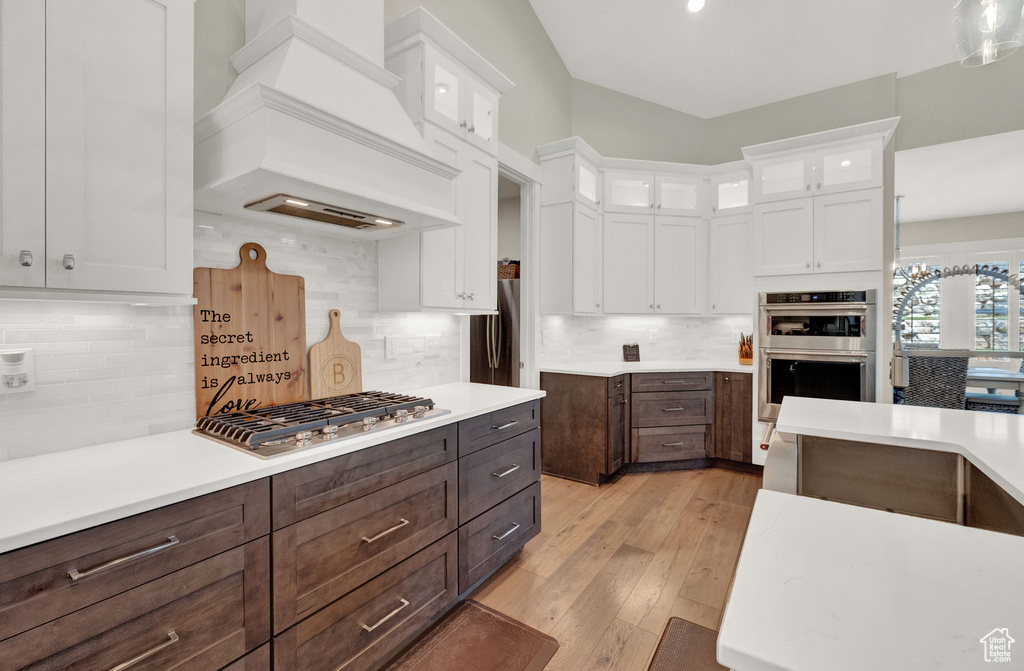 Kitchen featuring appliances with stainless steel finishes, white cabinets, backsplash, light wood-type flooring, and custom range hood