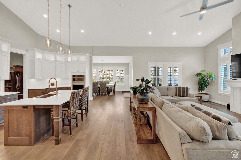 Living room with ceiling fan with notable chandelier, light wood-type flooring, sink, and high vaulted ceiling