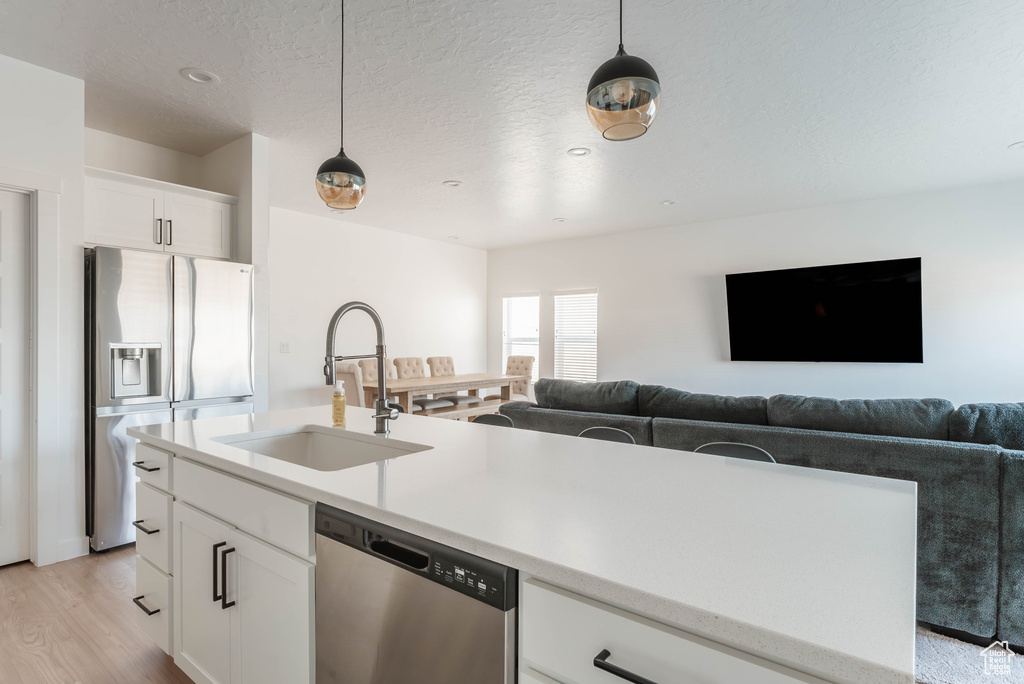 Kitchen featuring white cabinetry, stainless steel appliances, hanging light fixtures, light wood-type flooring, and a center island with sink