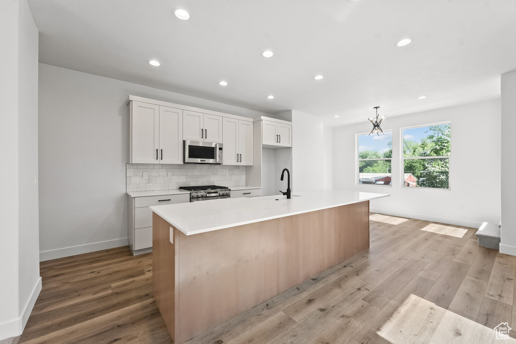 Kitchen with white cabinetry, appliances with stainless steel finishes, light hardwood / wood-style flooring, backsplash, and a kitchen island with sink