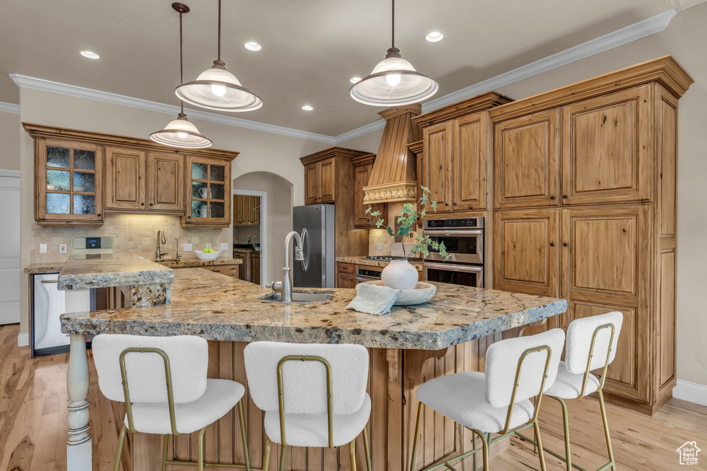 Kitchen featuring tasteful backsplash, stainless steel appliances, light wood-type flooring, a center island with sink, and custom range hood