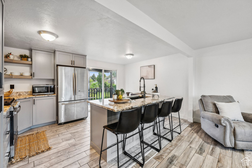 Kitchen with gray cabinets, light stone countertops, appliances with stainless steel finishes, light hardwood / wood-style flooring, and a kitchen island with sink