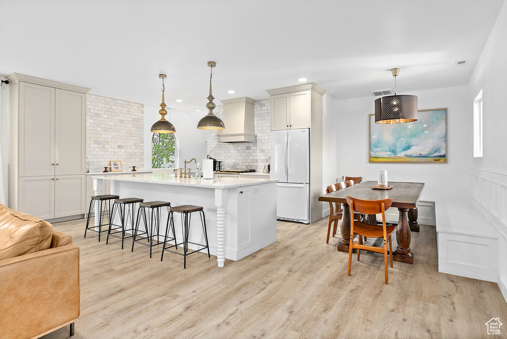 Kitchen featuring white fridge, tasteful backsplash, a center island with sink, custom range hood, and light hardwood / wood-style floors