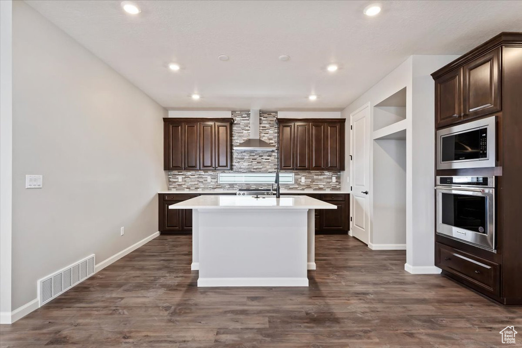 Kitchen with tasteful backsplash, wall chimney range hood, dark hardwood / wood-style flooring, an island with sink, and appliances with stainless steel finishes