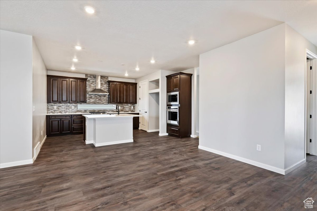 Kitchen featuring wall chimney range hood, dark hardwood / wood-style flooring, stainless steel appliances, decorative backsplash, and a center island with sink