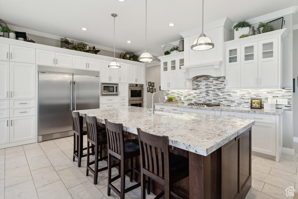 Kitchen featuring built in appliances, a center island with sink, backsplash, decorative light fixtures, and white cabinetry