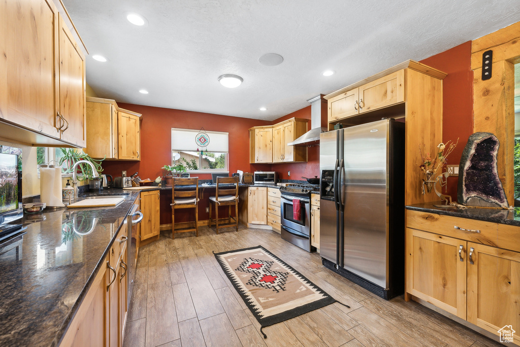 Kitchen with stainless steel appliances, wall chimney exhaust hood, sink, dark stone countertops, and light hardwood / wood-style flooring