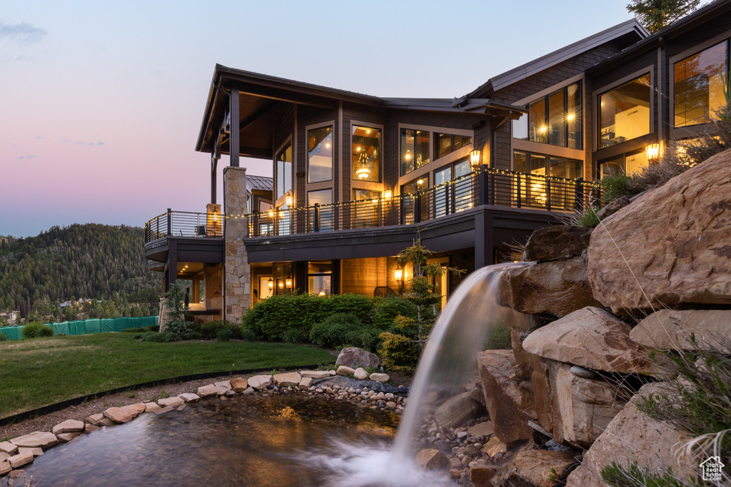 Back house at dusk with a mountain view, a balcony, and a lawn