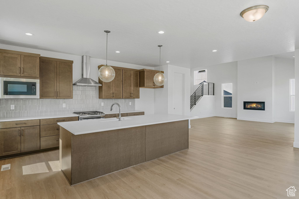 Kitchen with wall chimney range hood, stainless steel microwave, light hardwood / wood-style floors, decorative backsplash, and an island with sink