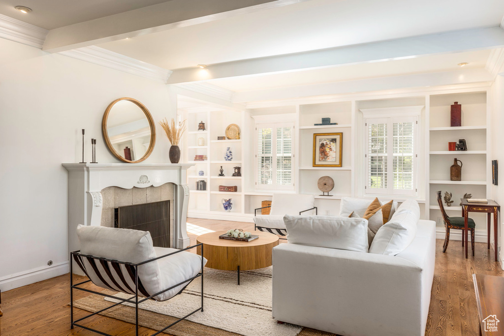 Living room featuring light wood-type flooring, built in shelves, ornamental molding, beam ceiling, and a tiled fireplace