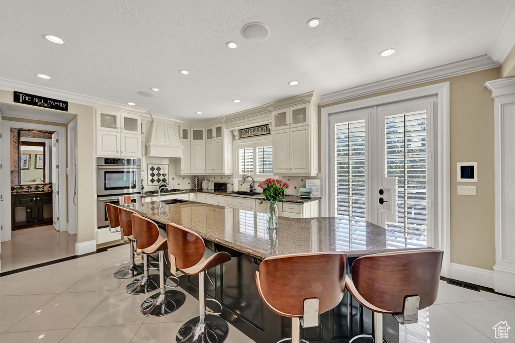 Kitchen with dark stone counters, light tile patterned flooring, stainless steel double oven, custom range hood, and a spacious island