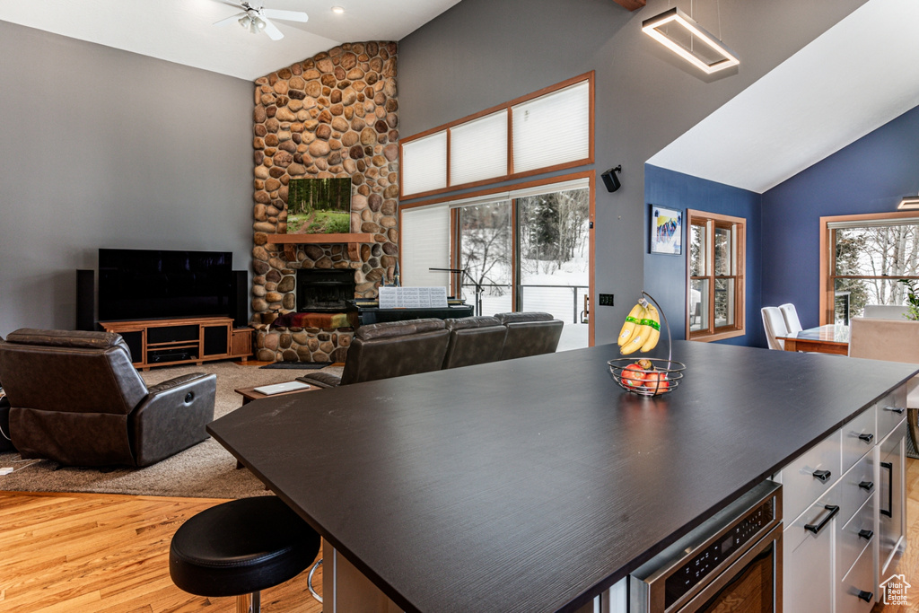 Kitchen featuring a stone fireplace, light wood-type flooring, a center island, ceiling fan, and high vaulted ceiling