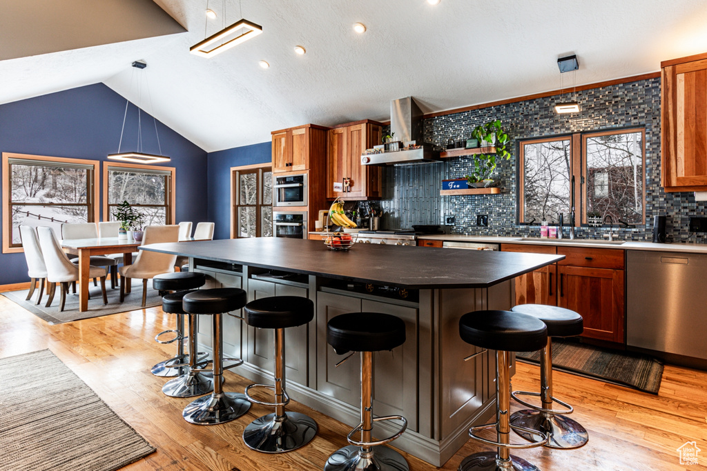 Kitchen with light hardwood / wood-style flooring, wall chimney range hood, backsplash, a kitchen island, and stainless steel appliances