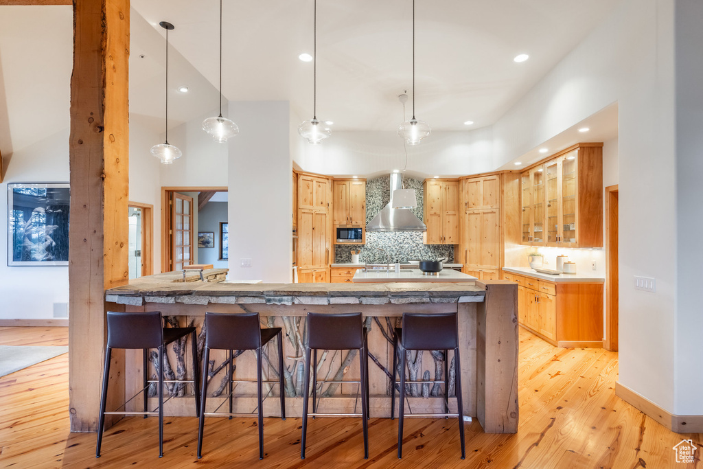 Kitchen with tasteful backsplash, light wood-type flooring, wall chimney exhaust hood, stainless steel microwave, and a kitchen bar