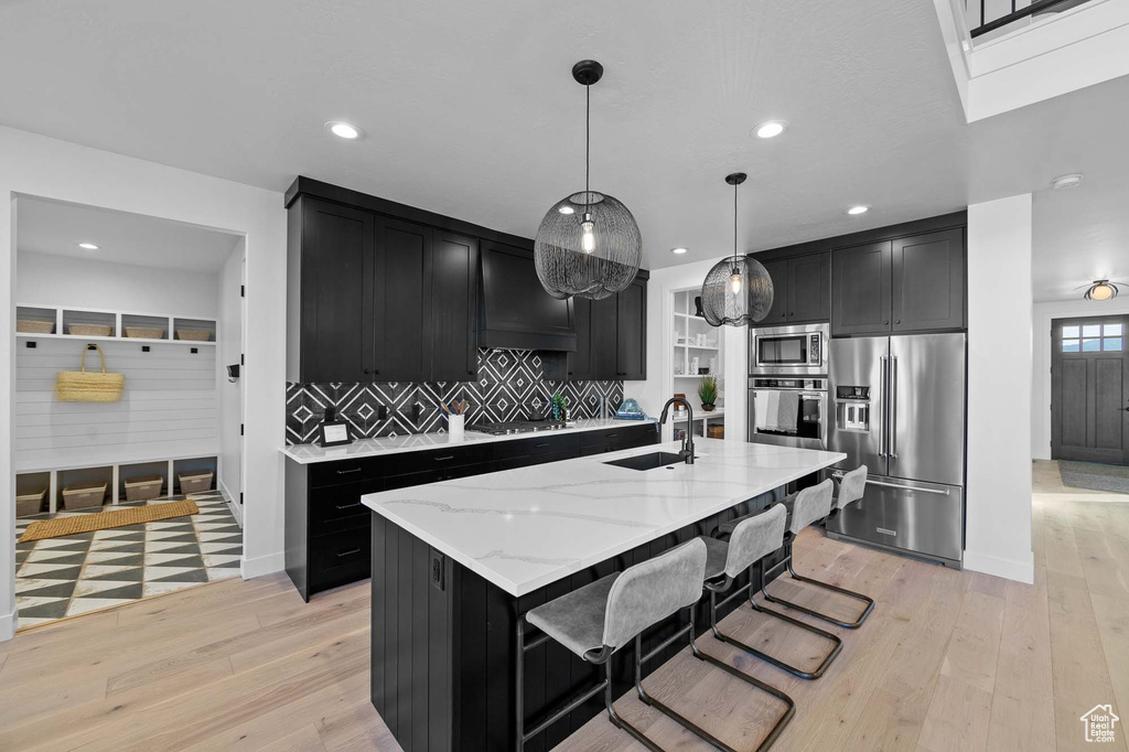 Kitchen featuring light stone counters, backsplash, an island with sink, appliances with stainless steel finishes, and pendant lighting