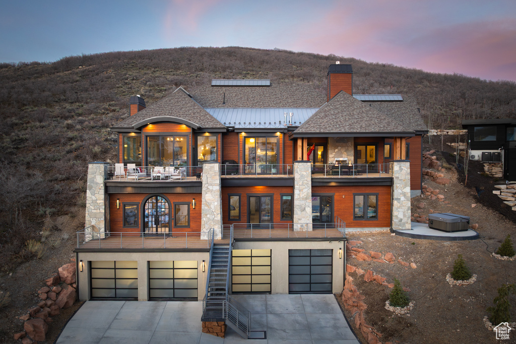 Back of property at dusk featuring concrete driveway, a chimney, an attached garage, and a balcony