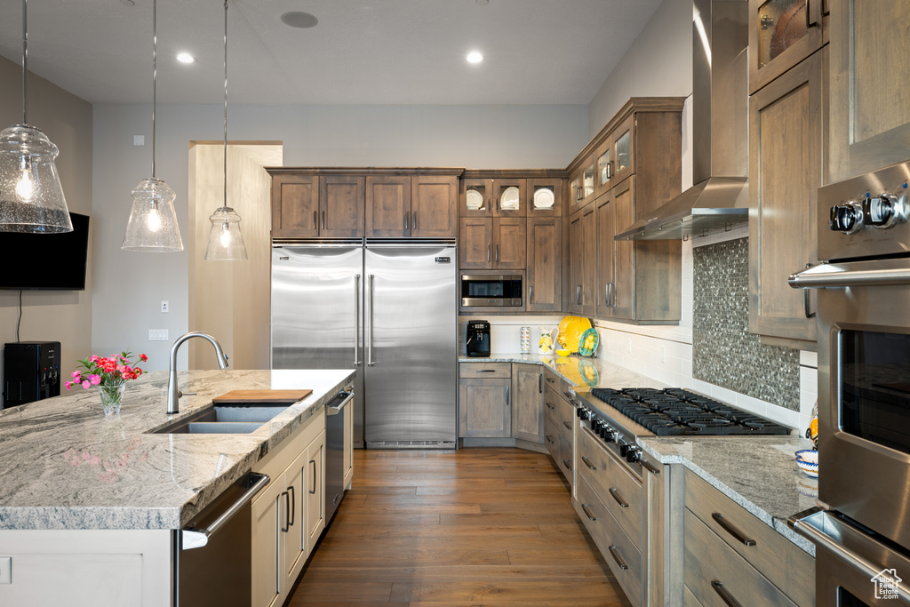 Kitchen with built in appliances, backsplash, light stone countertops, wall chimney exhaust hood, and a sink