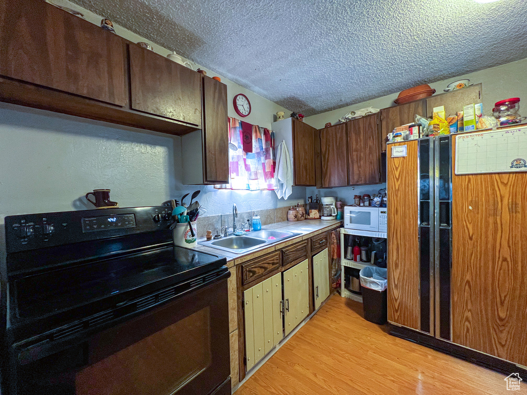 Kitchen with light hardwood / wood-style flooring, a textured ceiling, refrigerator, black / electric stove, and sink