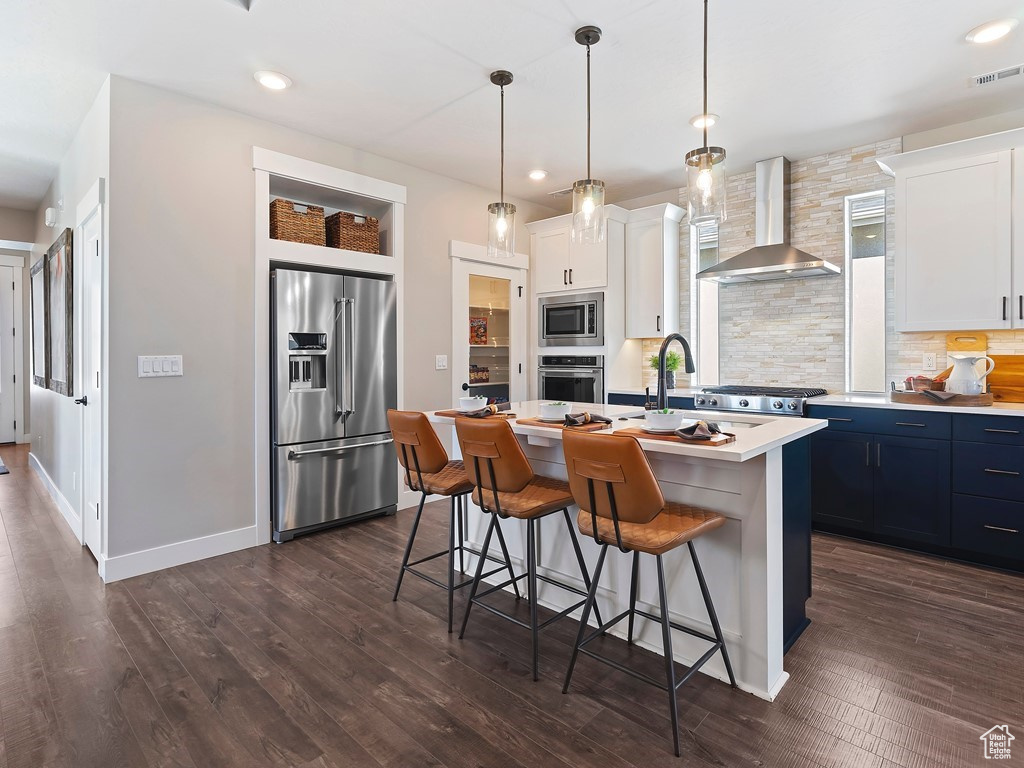 Kitchen with wall chimney range hood, dark hardwood / wood-style floors, appliances with stainless steel finishes, decorative light fixtures, and a center island with sink