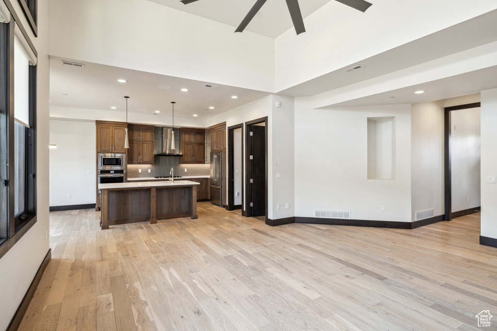 Kitchen featuring appliances with stainless steel finishes, wall chimney range hood, pendant lighting, a kitchen island with sink, and ceiling fan