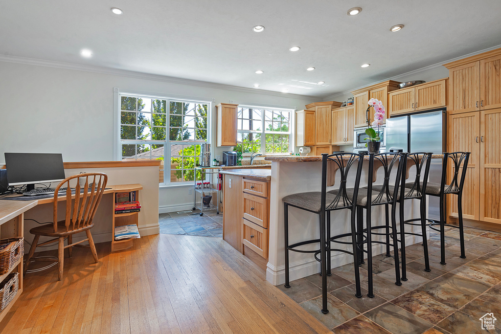 Kitchen featuring a breakfast bar area, appliances with stainless steel finishes, light brown cabinetry, hardwood / wood-style flooring, and ornamental molding