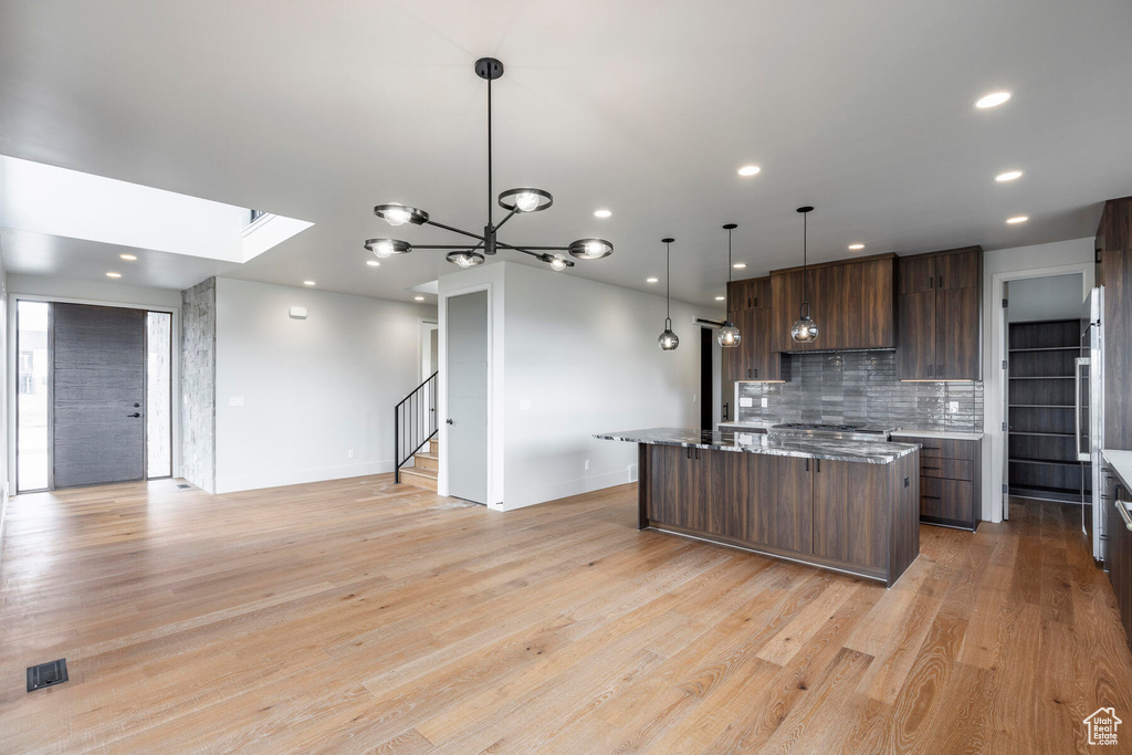 Kitchen with light stone countertops, decorative light fixtures, an island with sink, dark brown cabinets, and light wood-type flooring