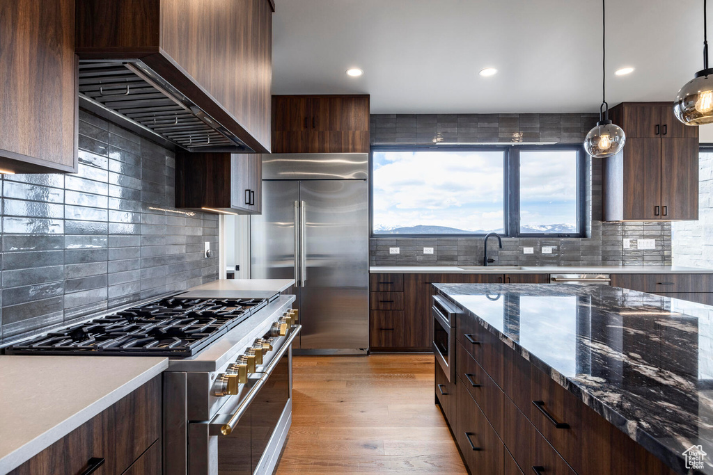 Kitchen with light hardwood / wood-style floors, tasteful backsplash, custom exhaust hood, premium appliances, and pendant lighting