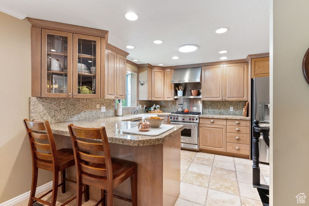 Kitchen featuring light tile patterned floors, premium stove, wall chimney exhaust hood, kitchen peninsula, and backsplash