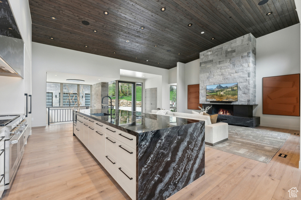 Kitchen with dark stone counters, a center island with sink, a stone fireplace, light wood-type flooring, and wooden ceiling