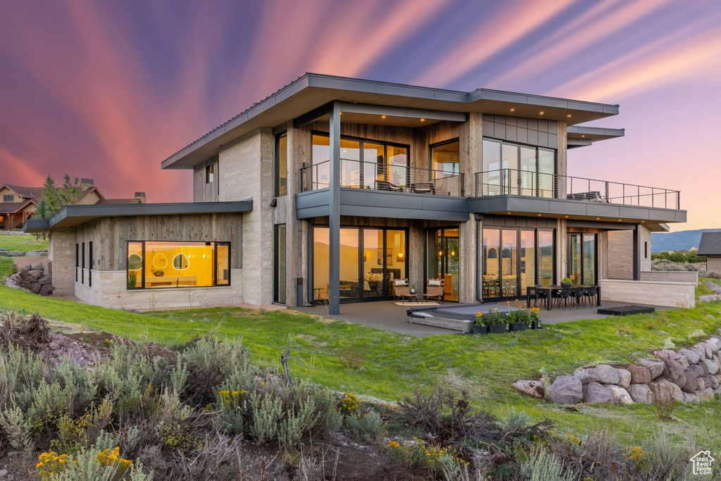 Back house at dusk featuring a balcony, a patio area, and a lawn